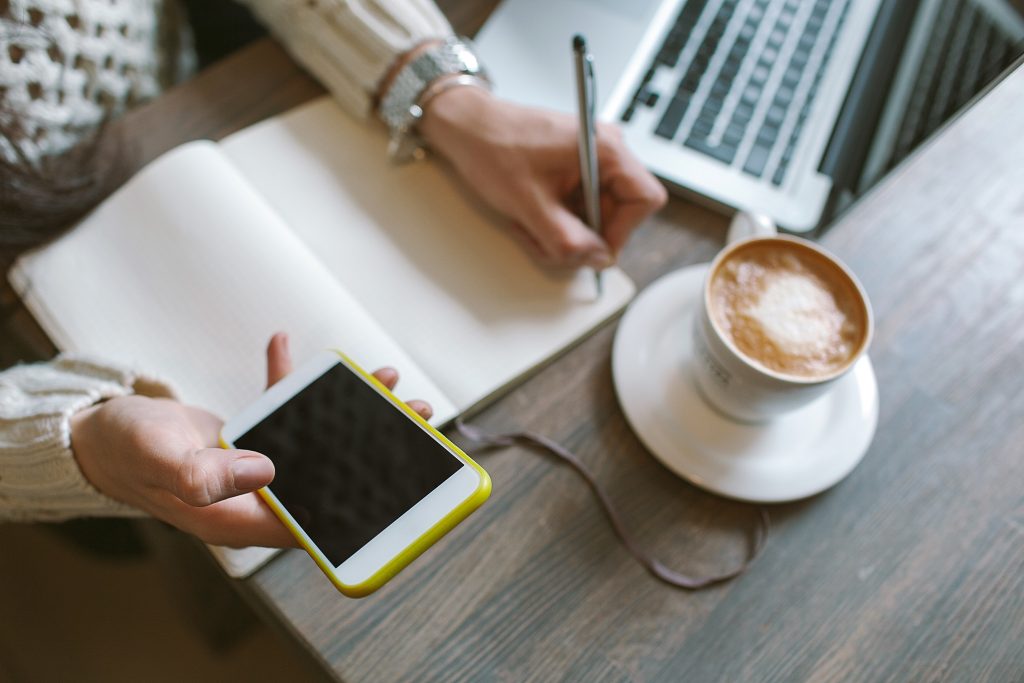 Hands of woman with pen and notepad, holding smartphone with cof